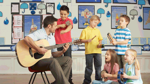 Male Teacher Playing Guitar With Pupils Having Music Lesson In Classroom